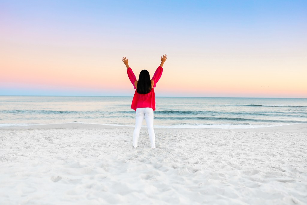 Person standing on the beach with arms raised, looking towards the ocean under a colorful sunset sky. Gulf Shores Alabama, Orange Beach, Alabama