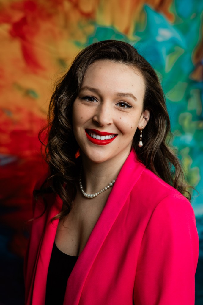 A smiling woman wearing a bright pink blazer and a pearl necklace poses in front of a vibrant, colorful background. Headshots in studio or on location for Fairhope, Gulf Shores, Orange Beach, Alabama.  For Real estate agents, corporate, commercial and education institutions.